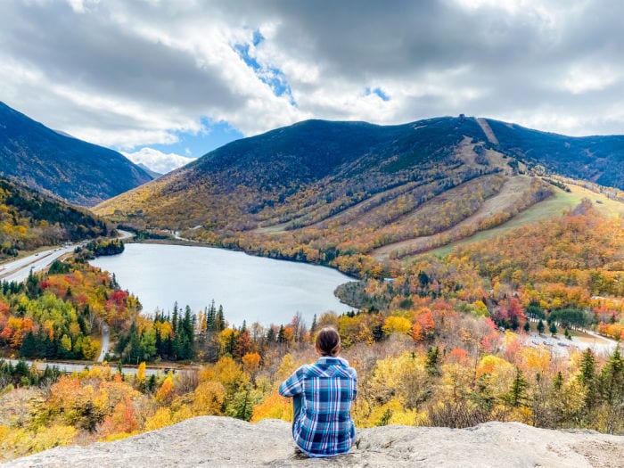 Woman sitting on Artists Bluff in NH overlooking Echo Lake with fall foliage on the trees and mountains
