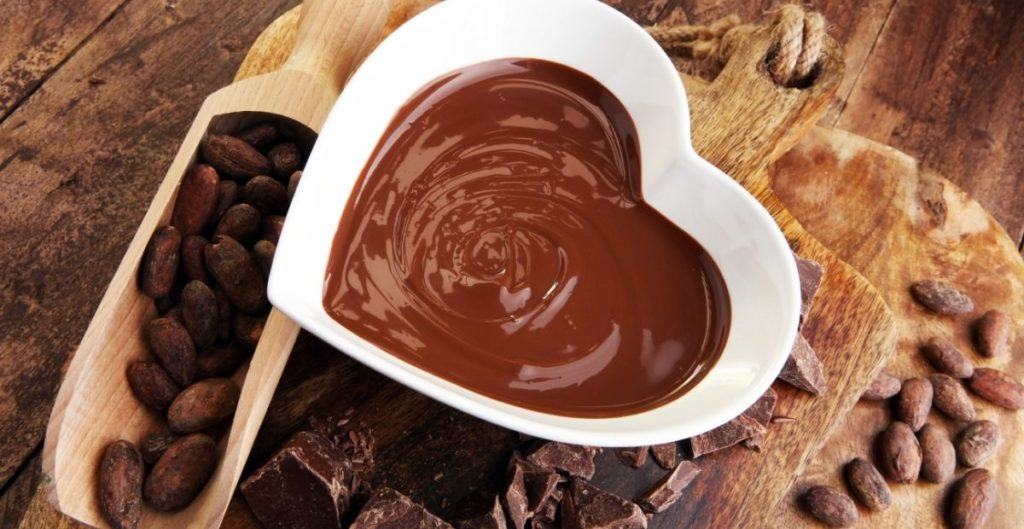 Melted chocolate in a heart bowl with pieces of chocolate and cacao beans surrounding on wood background