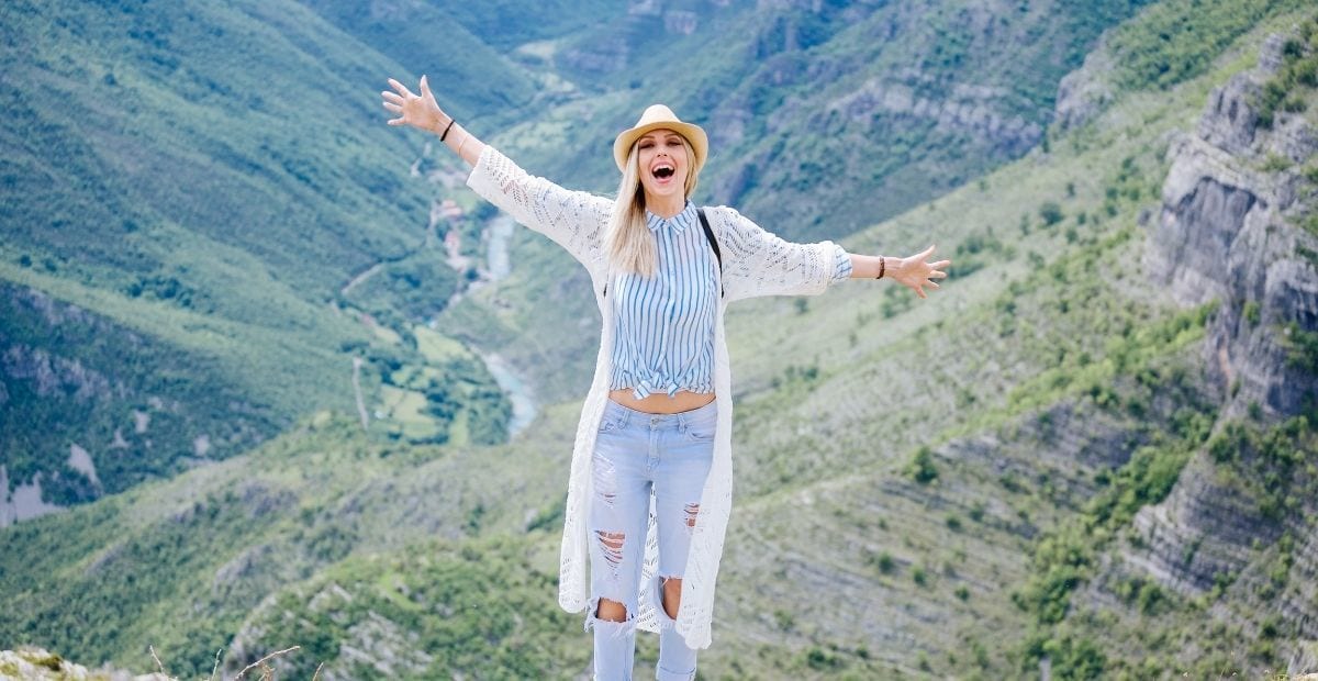 Teen girl in ripped jeans and a white sweater and hat with arms up in front of a valley