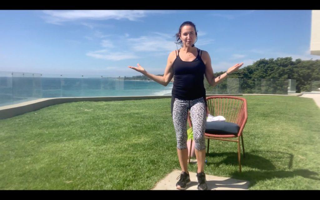 Woman on lawn in front of a beach in exercise clothes with a chair