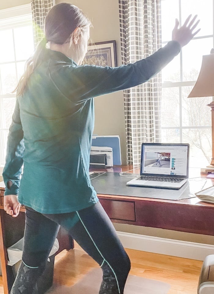 Woman in green shirt and black exercise pants exercising in front of desk with laptop showing exercise class