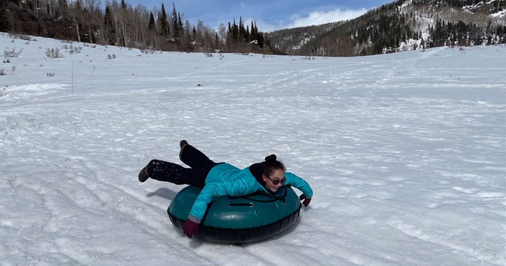 Girl in blue jacket and black snow pants laying on snow tube sliding down hill