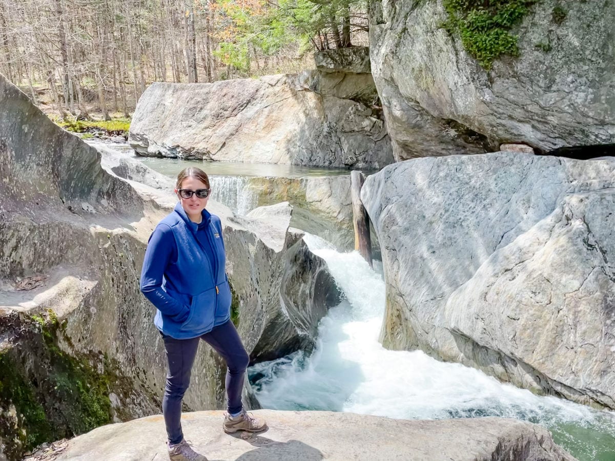 Woman in blue shirt and vest and black pants standing in front of waterfall and large rock boulders at Warren Falls Vermont