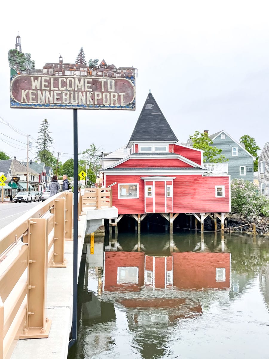 Welcome to Kennebunkport sign near bridge with red building and reflection in the water