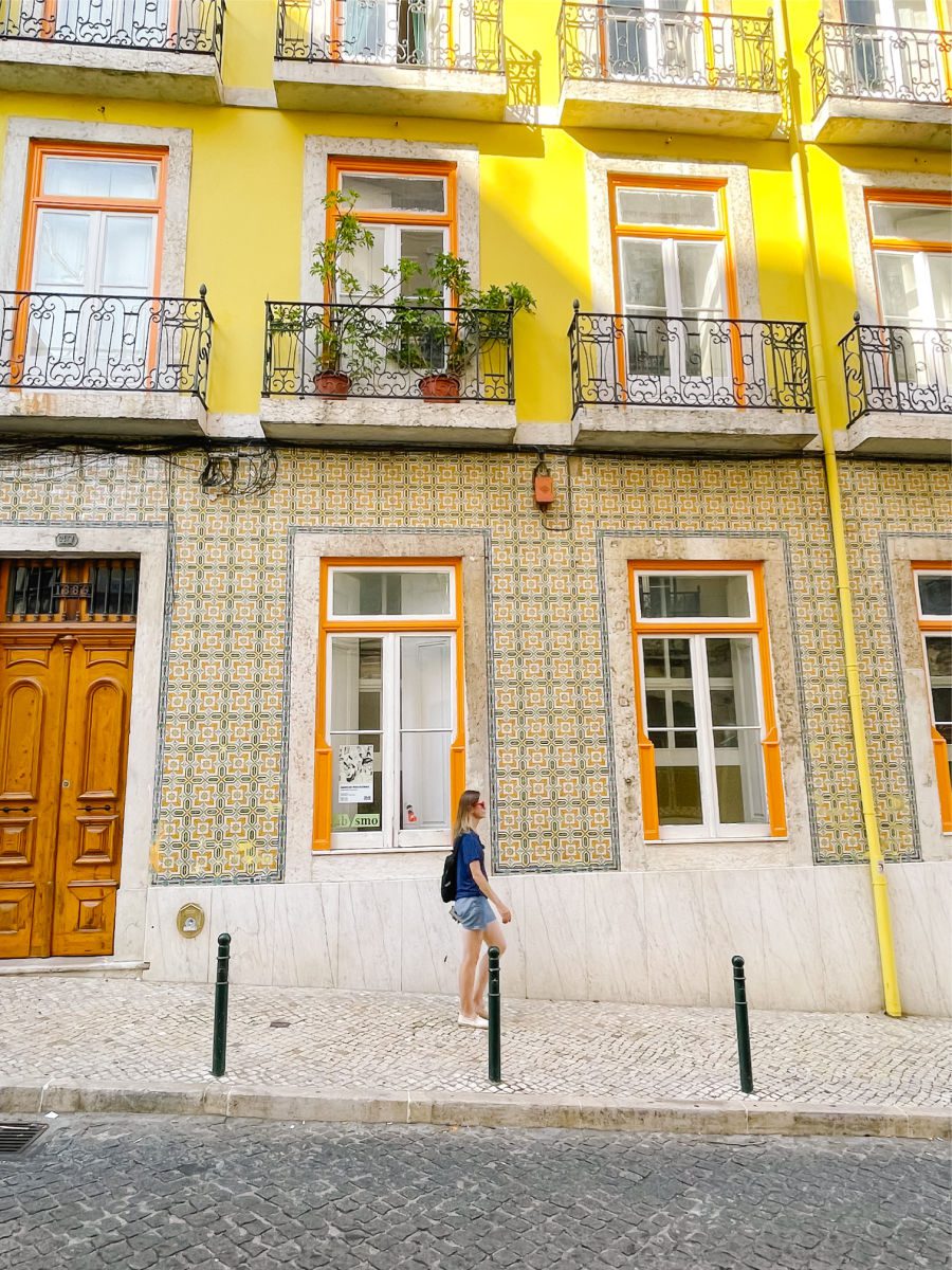 Woman in blue shirt and shorts walking past building with yellow tiles in Lisbon