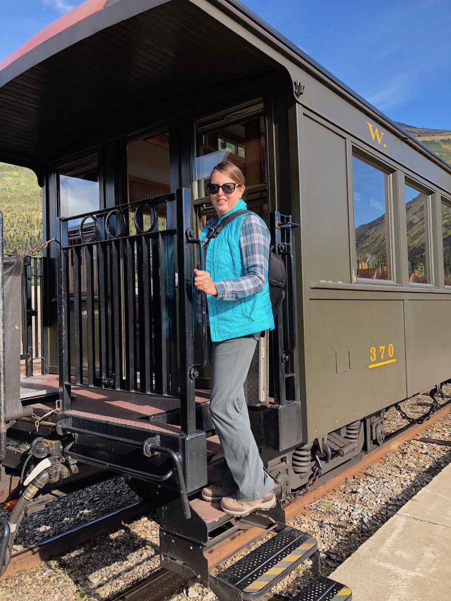 Tamara getting on train in turquoise vest, plaid shirt, grey pants and hiking boots