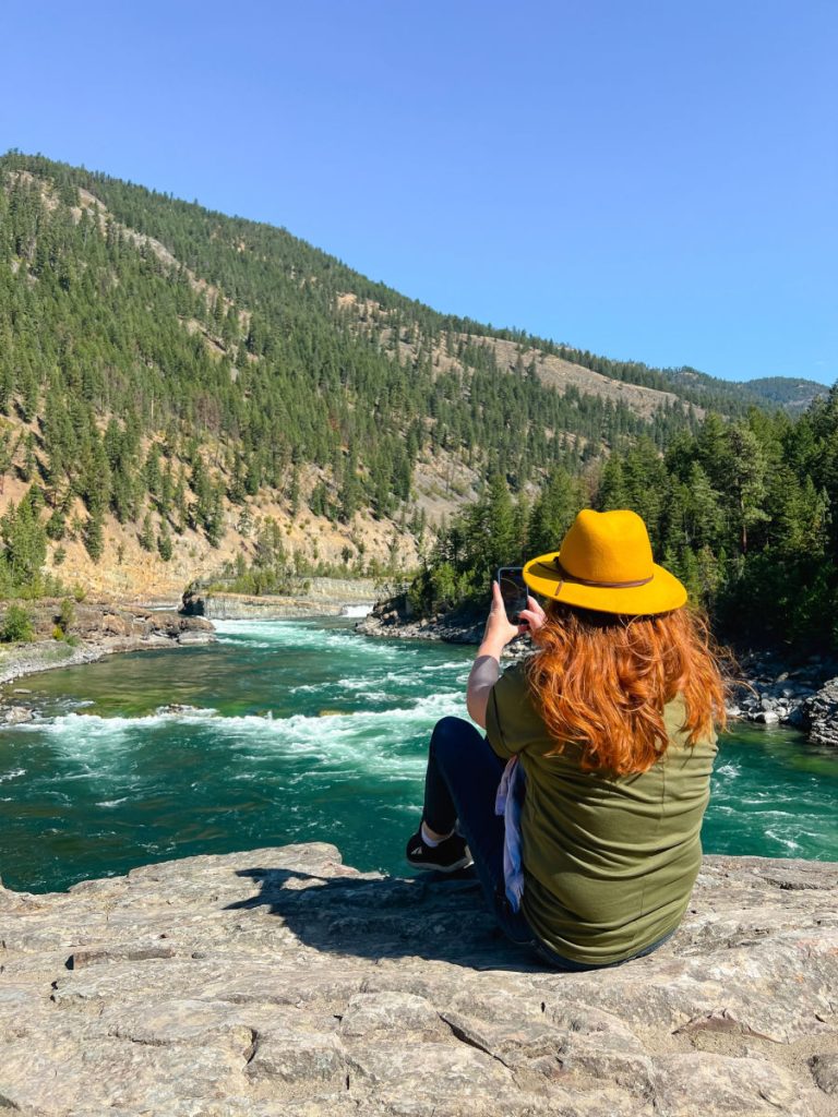 Keryn in green shirt and yellow hat taking a photo with her phone of the river while sitting on rocks above
