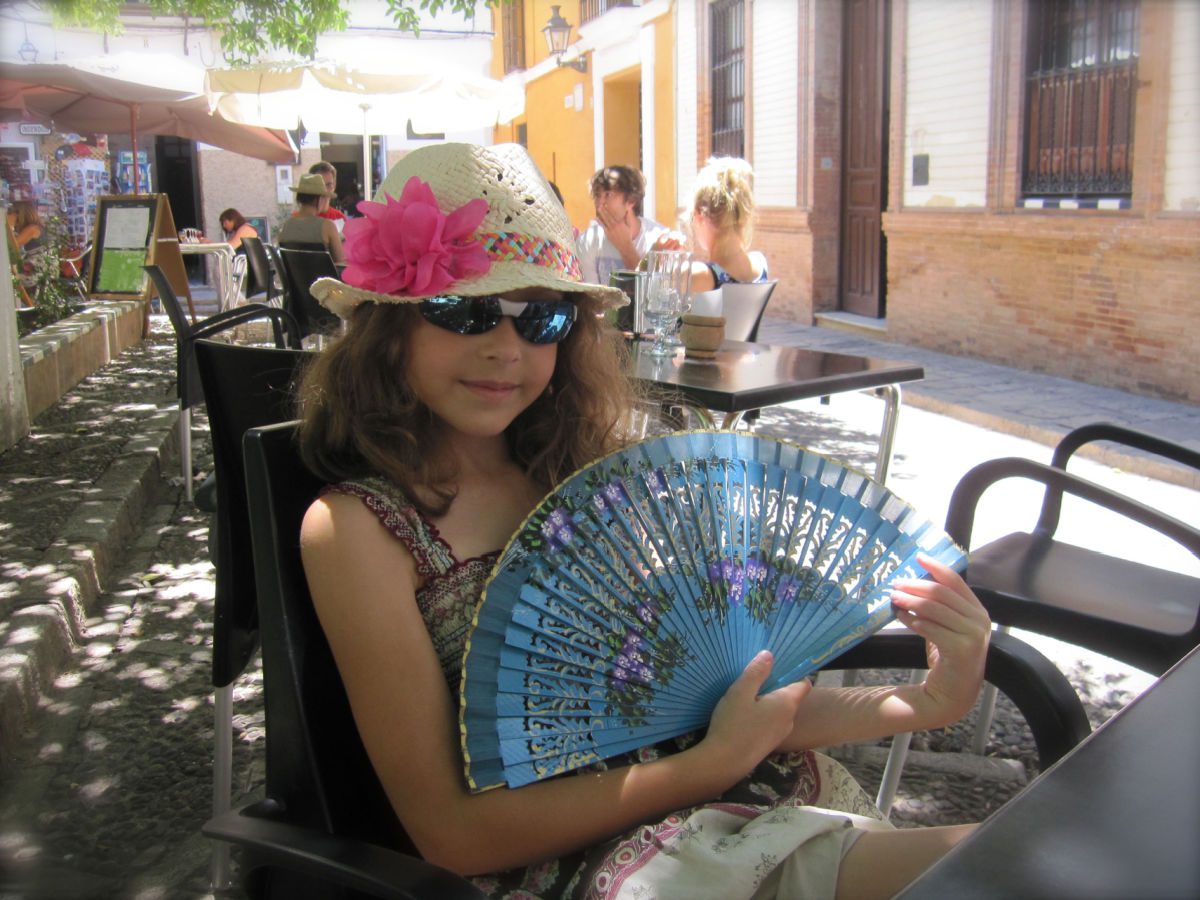 Girl with fan in hat and sunglasses at a table in a courtyard in Seville