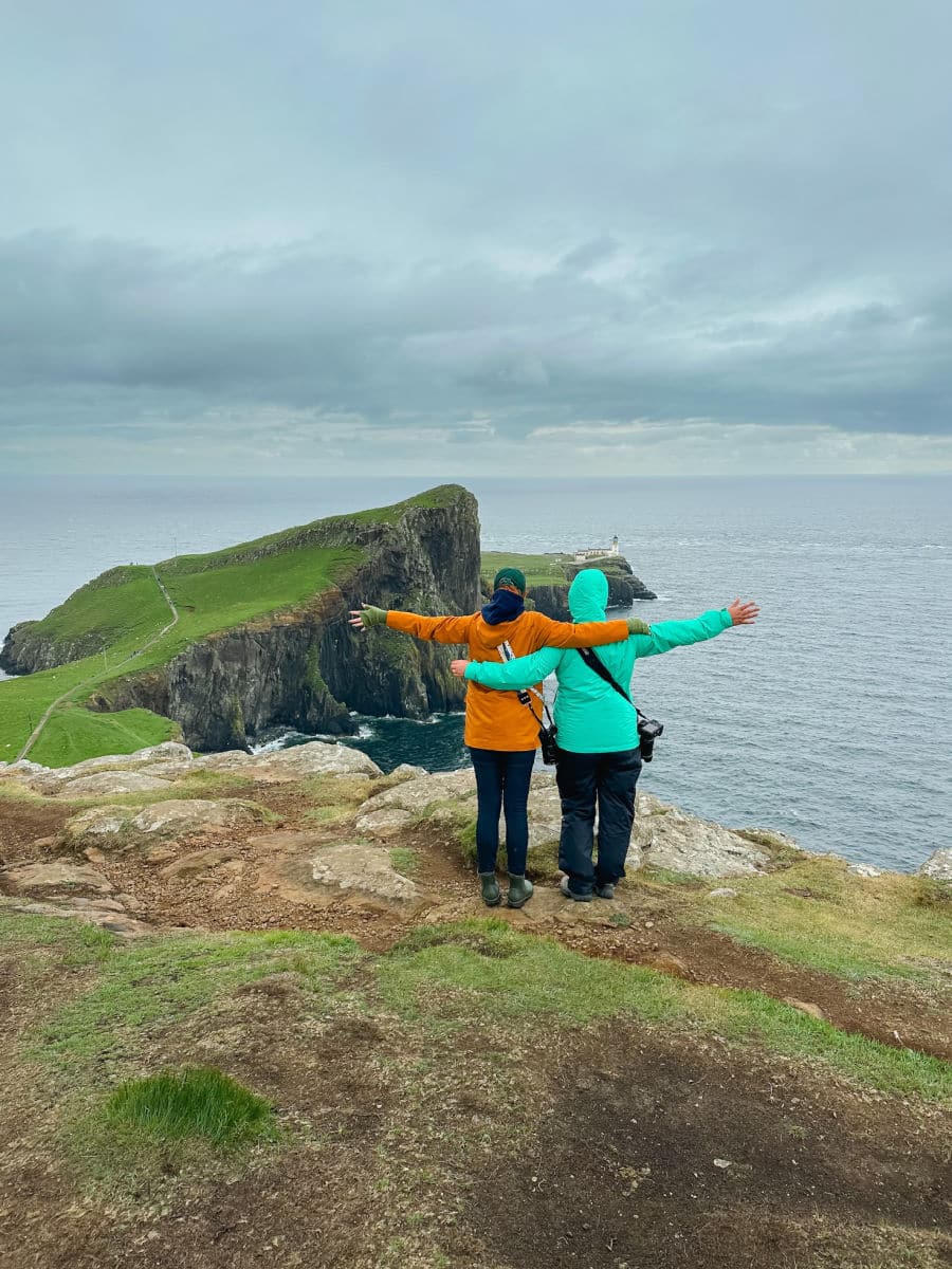 Keryn and Amanda in coats in and rain pants with arms out at Neist point